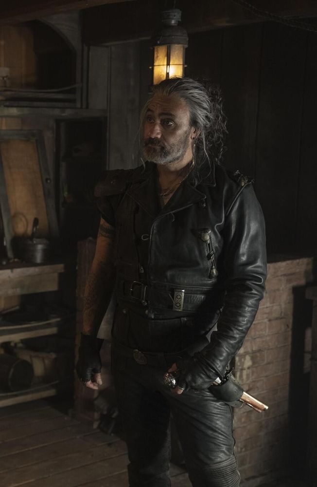 A picture from ofmd season 2 episode 2 of ed standing below deck in his leathers and slutty bun with a hand on his gun