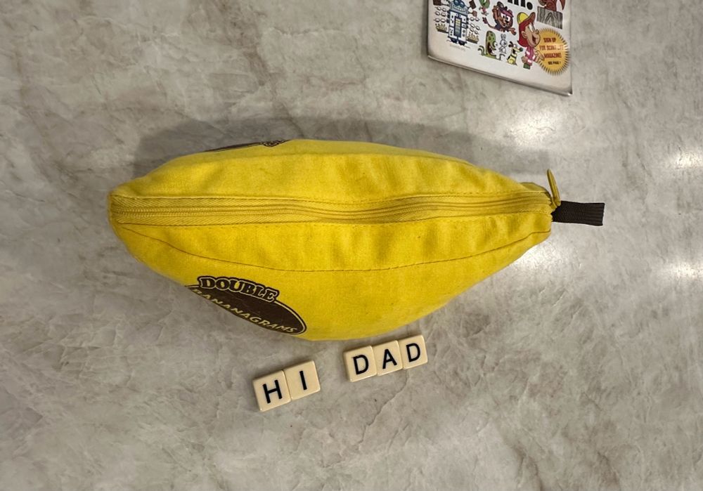 Bananagrams game in a yellow banana-shaped zipper pouch sits on a light quartz countertop. Below the pouch someone has used some letter tiles to spell “HI DAD.”