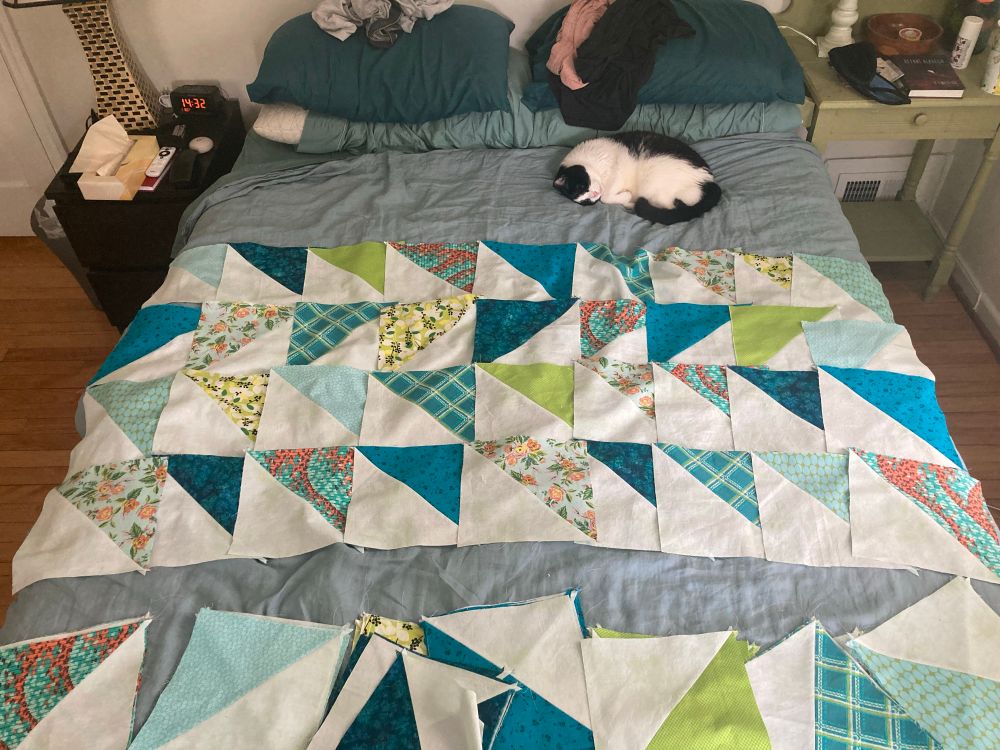 Tuxedo cat laying on top of a bed where I’m laying out quilt blocks
