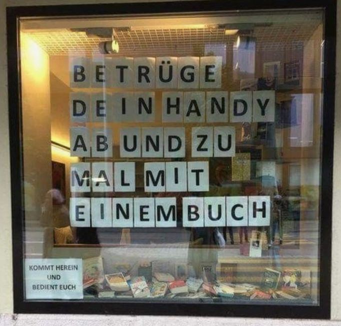 Das Bild zeigt das Schaufenster eines Buchladens. Im Fenster sind große Buchstaben angeordnet, die den Satz „BETRÜGE DEIN HANDY AB UND ZU MAL MIT EINEM BUCH“ Darunter steht ein kleineres Schild mit der Aufschrift „KOMMT HEREIN UND BEDIENET EUCH,“ 