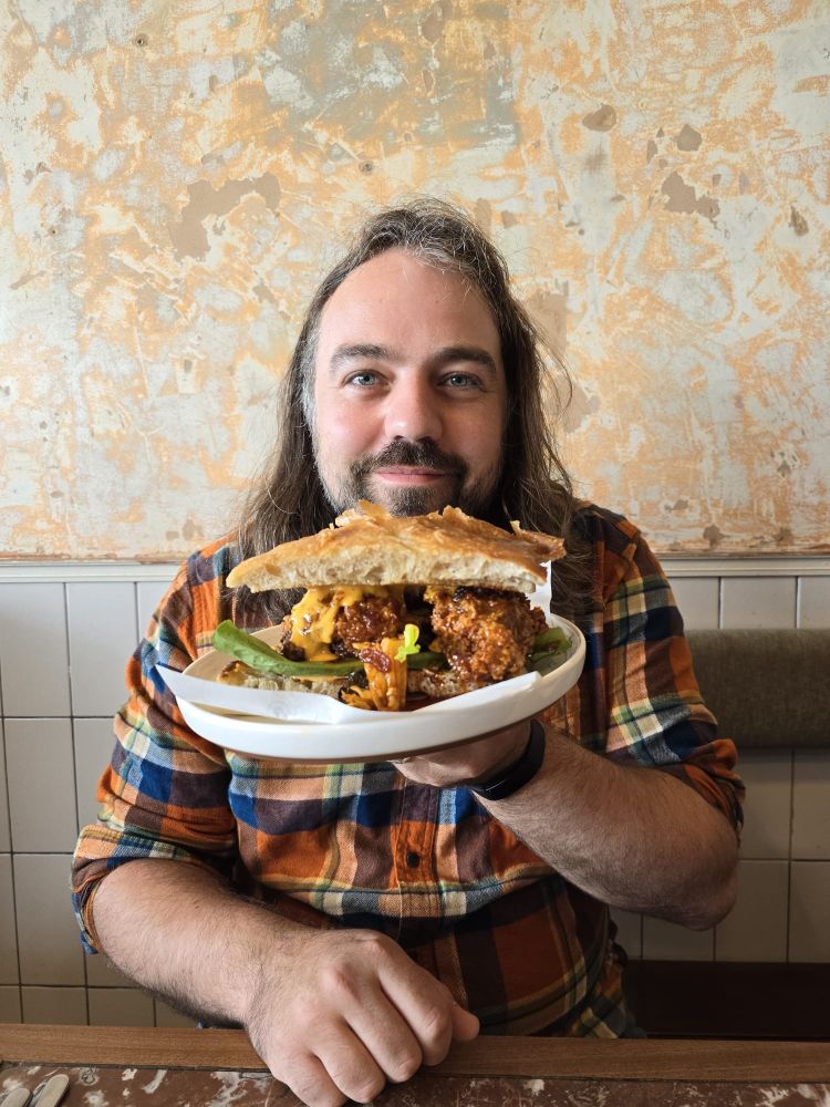 Paddy Freeland holding a big fried chicken sandwich in front of him in a plaid shirt sitting on a bench, over a marble table and a marble backdrop 