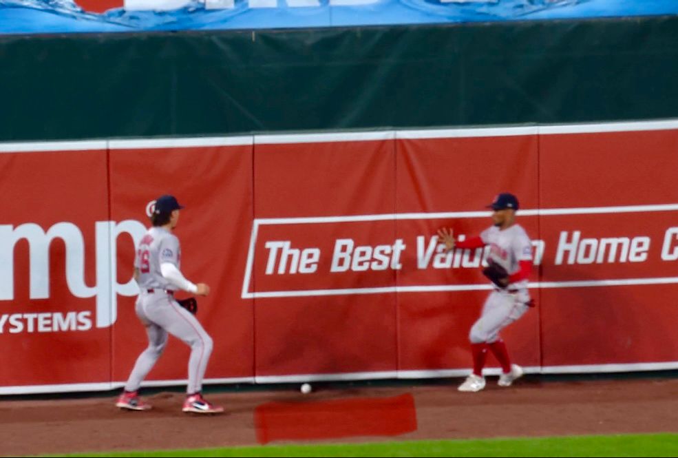 Two Red Sox outfielders stare at each other as the ball lodges beneath the padding on the wall