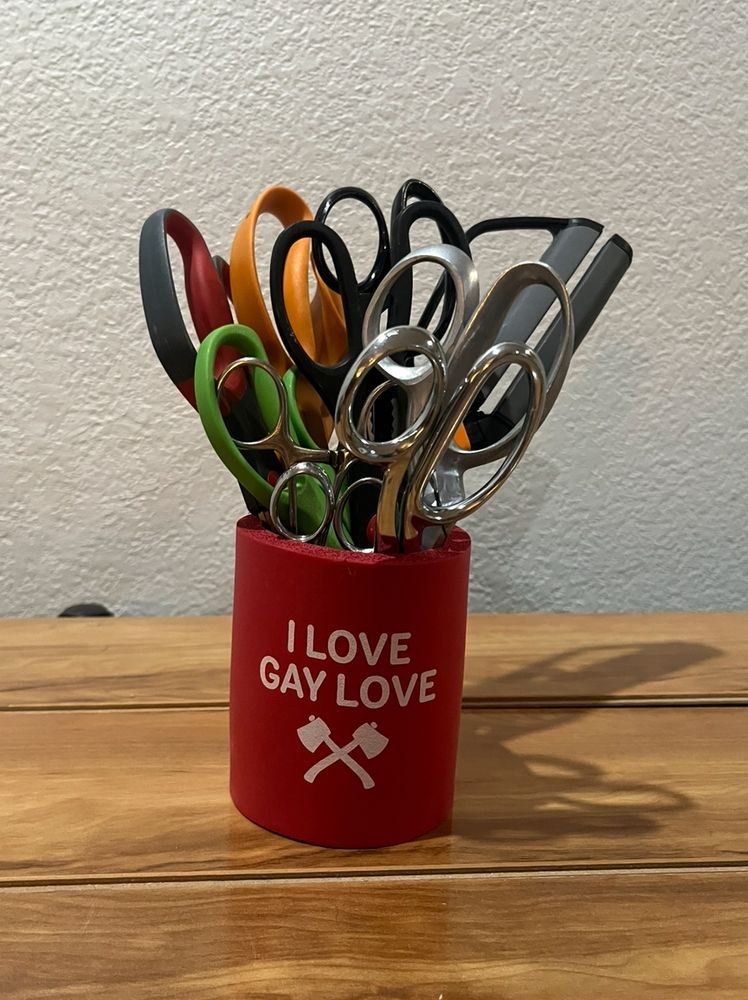 A dozen-plus pairs of scissors stored upright in a red cup emblazoned with I LOVE GAY LOVE and an illustrated image of two axes in love