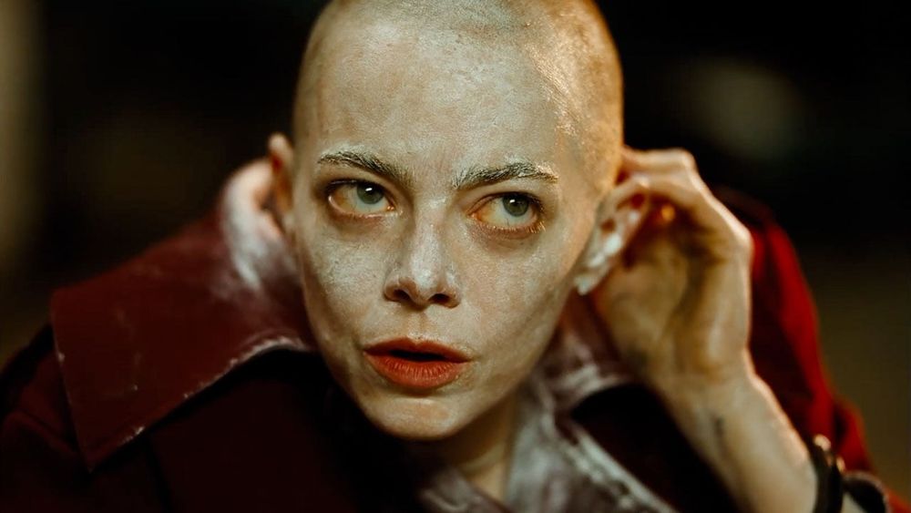 A bald Emma Stone playing captured CEO Michell Fuller, staring up at her captors, covered in white anti-rash cream, and dressed in a stained red overcoat. 