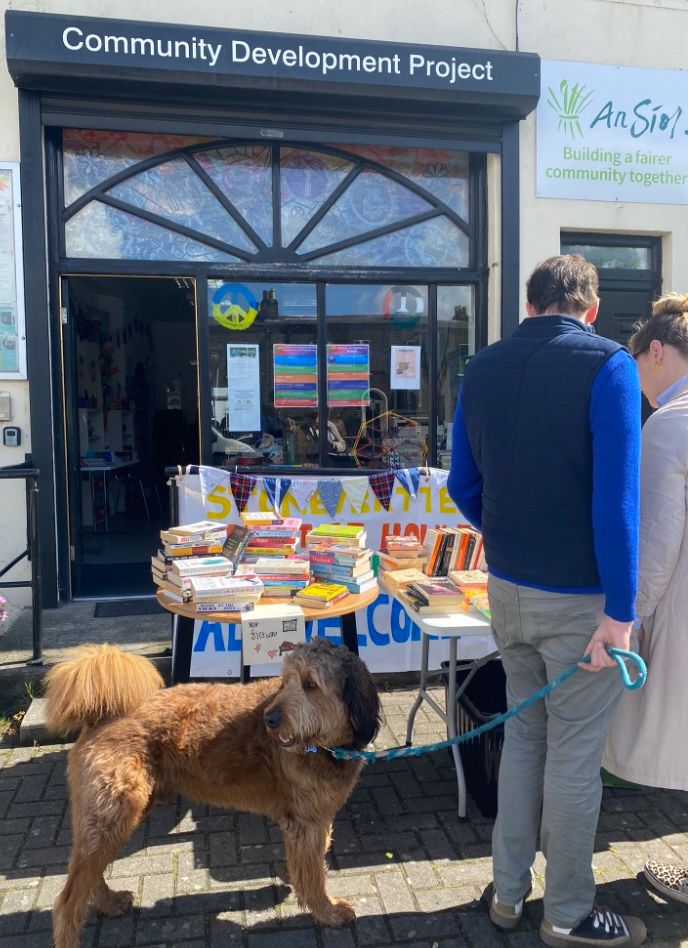 Tables covered in brightly coloured books on a sunny day. Two people are browsing with a gorgeous big brown dog.