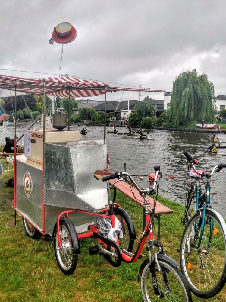Graue Bewölkung, grünes Flussufer, auf dem Fluss Kanupolospieler, im Vordergrund Blick auf ein rotes e-Dreirad, das einen selbstgebauten stahlfarbenen kleinen Auflieger zieht, auf dem sich eine Italien. Espressosiebträgermaschine in beige befindet, geschützt von einem rot-weiss-gestreuften Segeltuchdach und gekrönt von einem übergrossen Ausschnitt einer gefüllten roten Cappuccinotasse und -untertasse