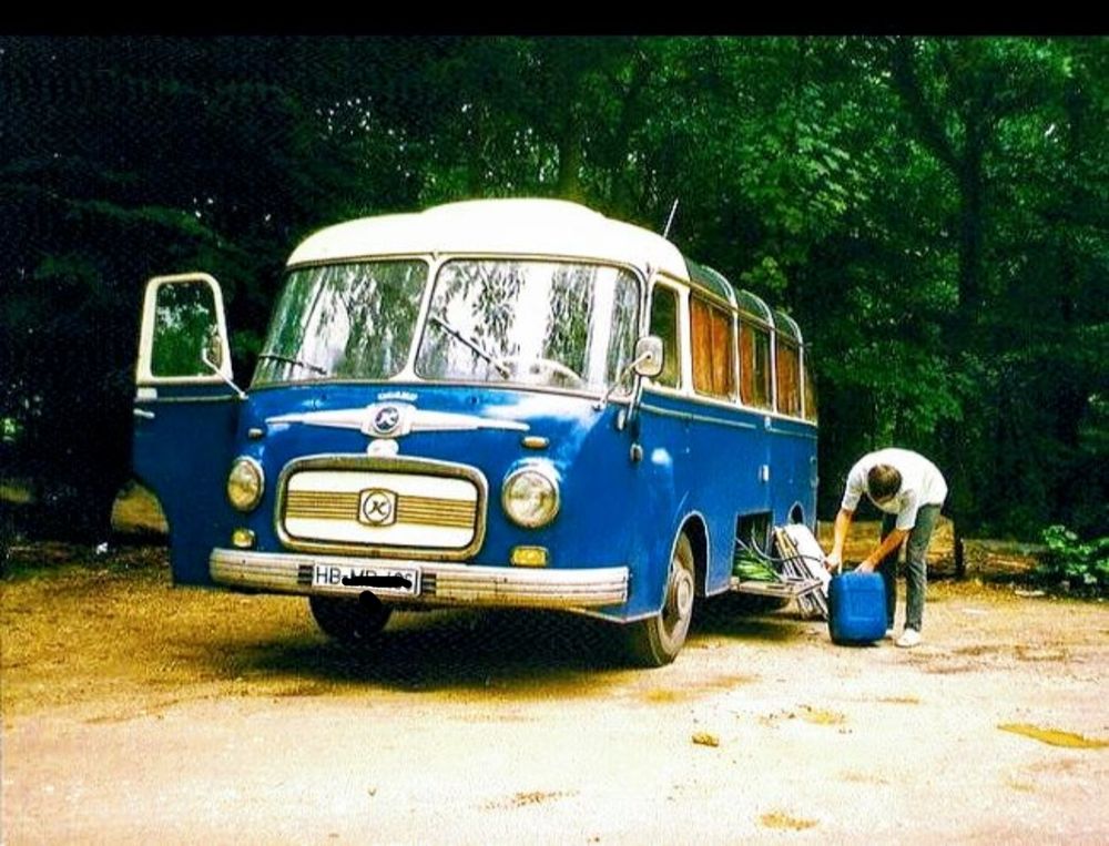 Blauer Oldtimerbus mit abgerundeten Kanten, weißem Dach und gewölbten Dachfenstern, auf einem sandigen Waldparkplatz mit sattem, dunkelgrünem Wald im Hintergrund und einem Menschen, der etwas aus einem ebenso blauen 10-Wasserkanister abfüllt