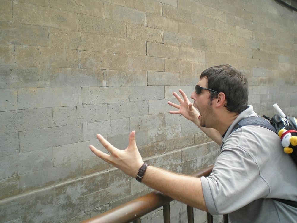 A man in sunglasses and a tan shirt yells at a brick wall. His arms are up in frustration. 