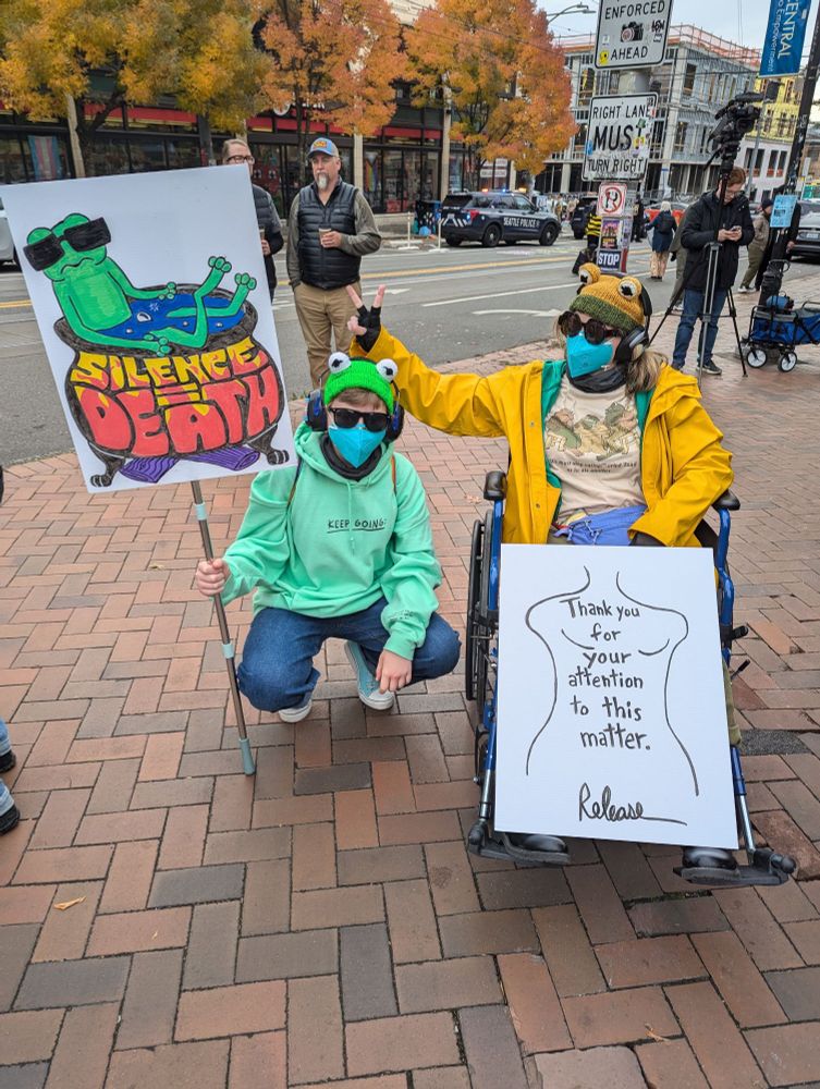 Two protestors with frog hats holding signs that say Silence = Death with a frog in a boiling pot of water on the left and "Thank you for your attention to this matter — Release" ('Release' as DJT's signature). The person on the left has sunglasses and a mask on wearing a green hoodie that says XL"Keep going". The person on the right is sitting in a wheel chair, is putting bunny ears above the person's head next to her, and is wearing a yellow rain coat and a Frog and Toad shirt. There are trees in the background across the street from the plaza with paved bricks the two people are in. There is a person with a video camera on a tripod in the background on the right.