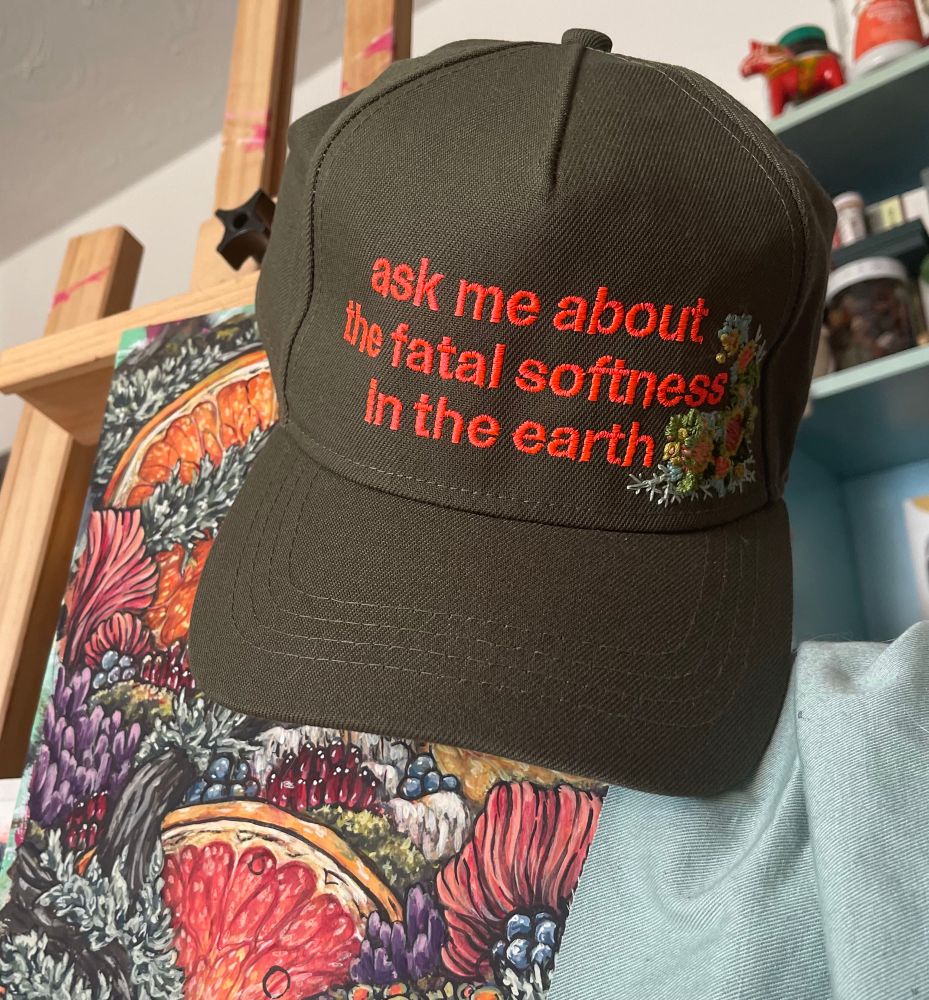 Olive green hat with the phrase ‘ask me about the fatal softness in the earth’ embroidered in coral thread hanging from a wooden easel