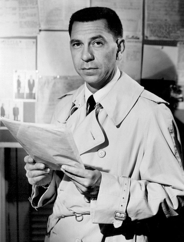 Portrait of actor Jack Webb as Detective Joe Friday from the early TV police procedural, Dragnet. White man with dark hair, conservatively groomed, wearing a suit and trench coat, holding file papers. 
