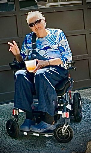 Picture of me, an older white lady with olive skin, white hair, wearing blue, sitting in my power chair in front of my house. 