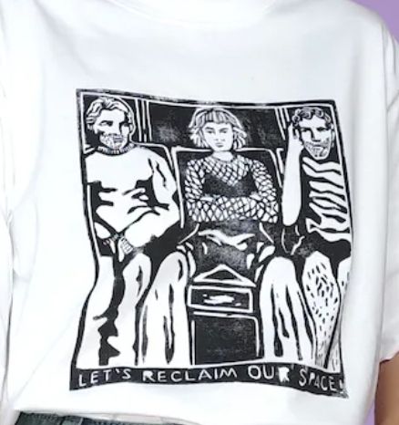 ein weißes t shirt mit einem schwarzen druck, auf den eine junge frau mit pony und netzoberteil breitbeinig in der bahn sitzt, neben ihr zwei komische dudes