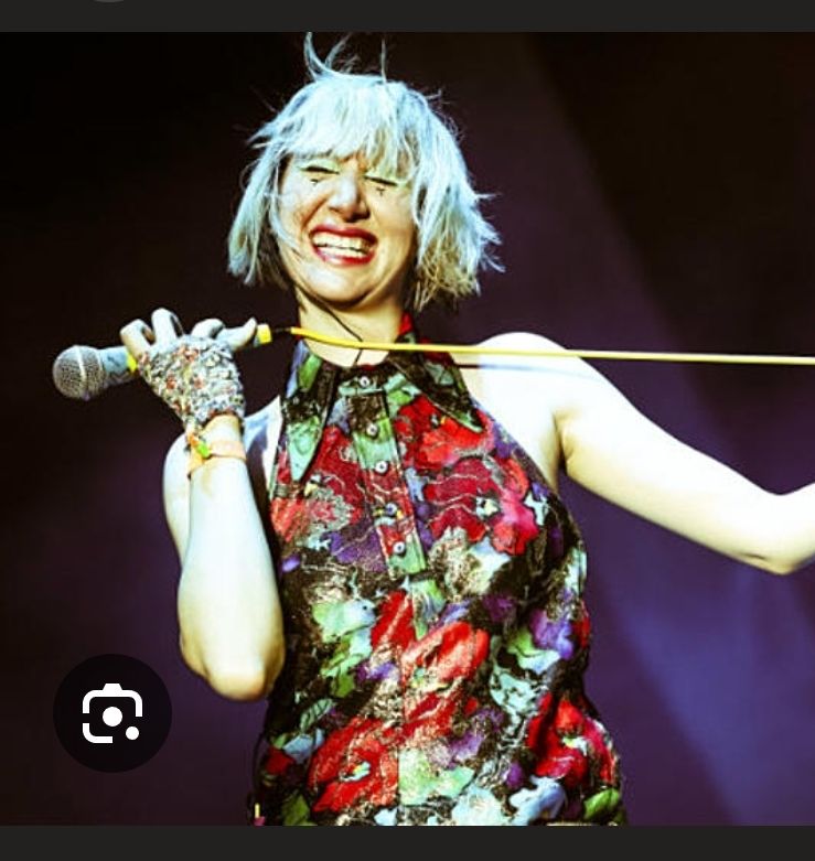 karen o in einem hochgeschlossenen ärmellosen lederoberteil wild gemustert in rot grün und schwarz lila, es hat einen spitzen kragen und knöpfe