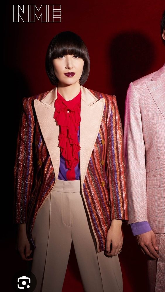 karen o in einer beigen anzughose mit bügelfalte, einem roten rüschenhemd und einem bunten glitzernden langen zirkusdirektorinnen blazer 