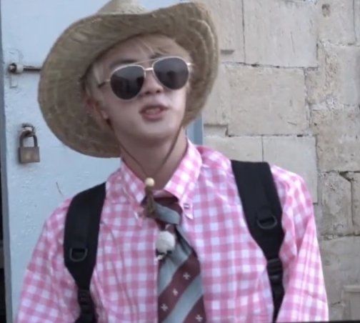 BTS Jin in Malta with a straw hat, pink gingham shirt & striped tie. 