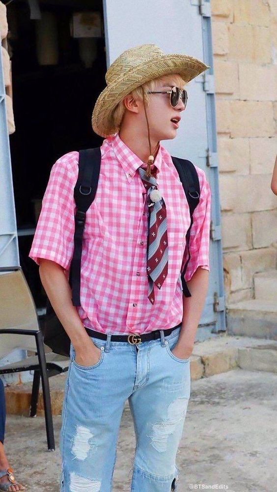 BTS Jin in Malta with a straw hat, jean shorts, pink gingham short sleeve shirt & tie. 