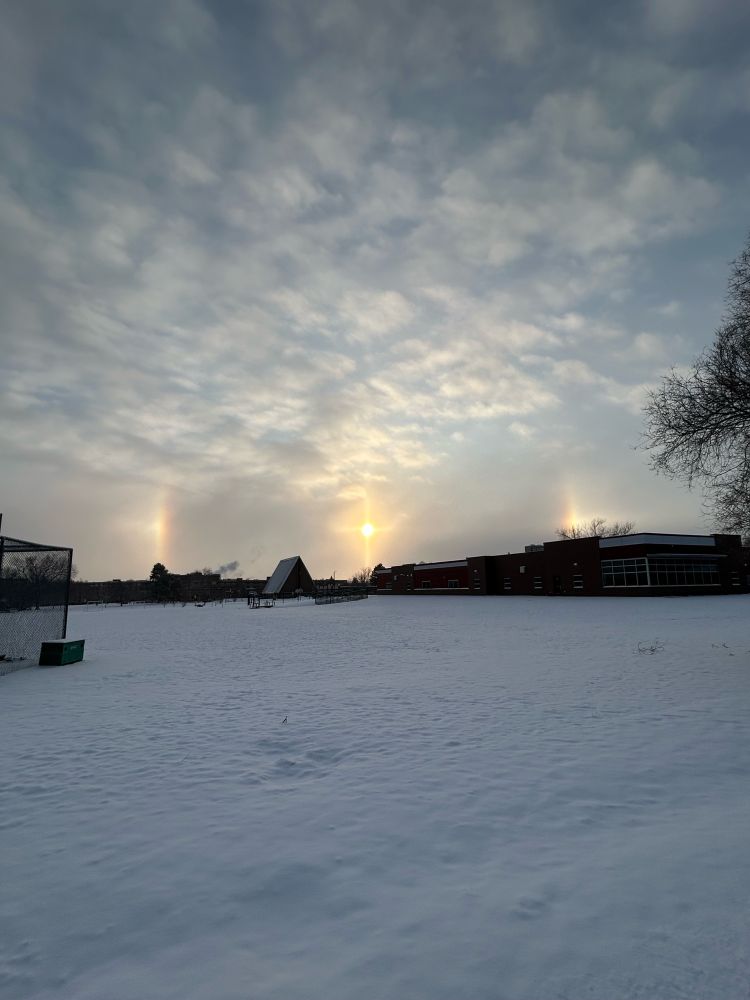 The sun rises over a snowy field. A bright circle of light - a sun dog - centers the sun.
