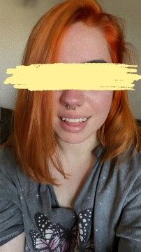 A person with bright orange shoulder-length hair is smiling at the camera. They have a septum piercing and are wearing a grey shirt with butterfly designs. Their eyes are covered with a yellow scribble for anonymity. The lighting is soft and natural, and the background appears to be indoors.