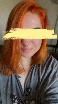The same person with bright orange hair is smiling slightly with a closed mouth. They are again wearing the same grey shirt with butterfly designs, and their septum piercing is visible. Their eyes are covered with a yellow scribble. The lighting and background are consistent with the first image, suggesting both were taken in the same setting.
