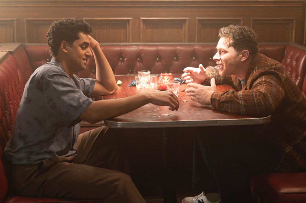 ravi and buck sitting in booth at bar, buck yapping with open waving hands and ravi is reaching for another drink and looking annoyed