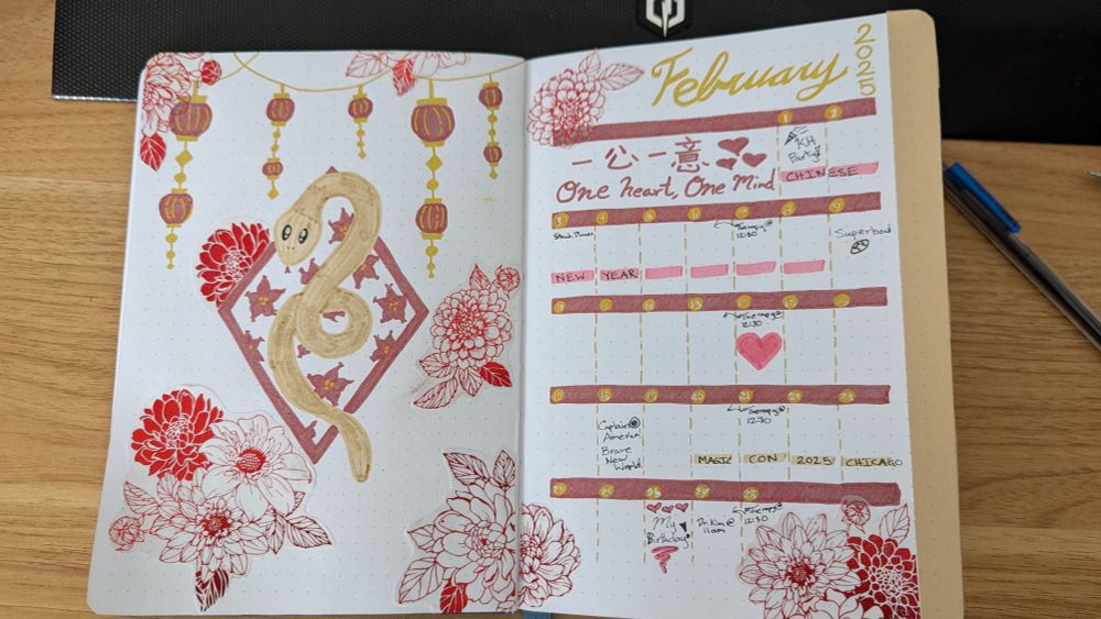 February Bujo page with a Chinese Year of the Snake New Year Theme. Left page shows a cute snake in a diamond of flowers. Above hangs small red lanterns. The right page shows the February 2025 calendar with One Heart, One Mind quote written in Chinese. The colors are deep red and gold. You can also see scatters of red flowers everywhere cut from a vellum origami paper.
