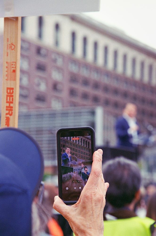 Color photo of a person recording Rep. Brendan Boyle speaking. Boyle is on the screen and out of focus in the background.
