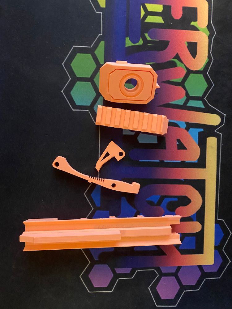Collection of pale orange 3d printed parts for a string powered dart blaster. There is a square muzzle, a trigger, a small section of picatinny rail, a large rectangular bar with raised center, and a lever with cuts made to imitate a spring action.