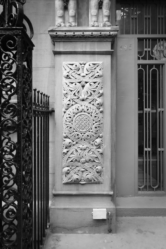 #ClassicMono photo of Romanesque relief panel in an apartment building facade, framed by various styles of ironwork.