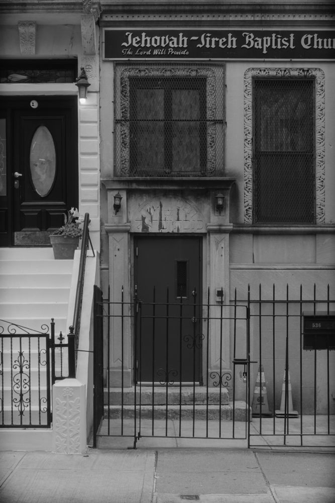 Monochrome photo of a street view on West 148th street. Details abound in gates and grates, stained glass and fire cones, incised relief Hebrew lettering and a Baptist church sign. I love this city.