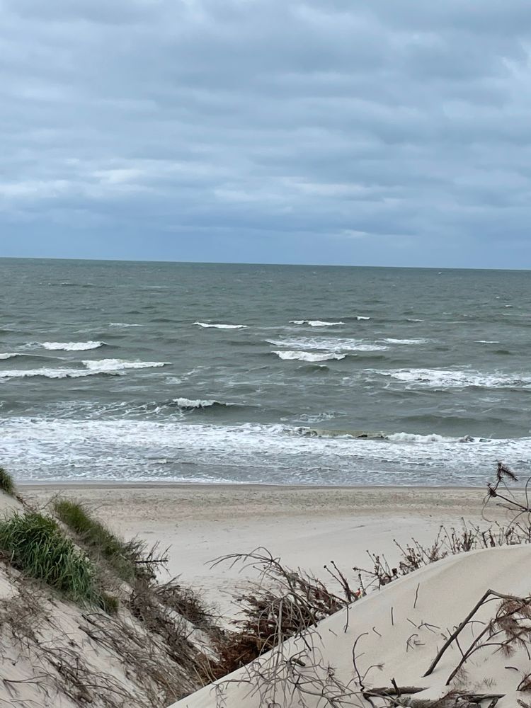Die umgewühlte, leicht graue Nordsee aus den Dünen heraus fotografiert. Zwei Dünen geben einen V-förmlichen Einblick auf den Strand und das Meer frei 