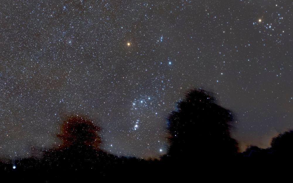 Aikavalotuksella otettu kuva talvisesta tähtitaivaasta etelän suuntaan. Puiden siluetit kuvan alaosassa. Keskellä Orionin tähdistö, vasemmalla alhaalla puiden välistä näkyy Sirius. Härrän tähdistö yläoikealla.