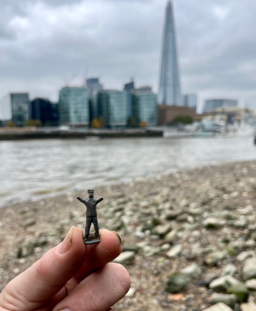 A small figurine of a person with their arms raised. In the background are the gravels of the foreshore and the Shard in the distance