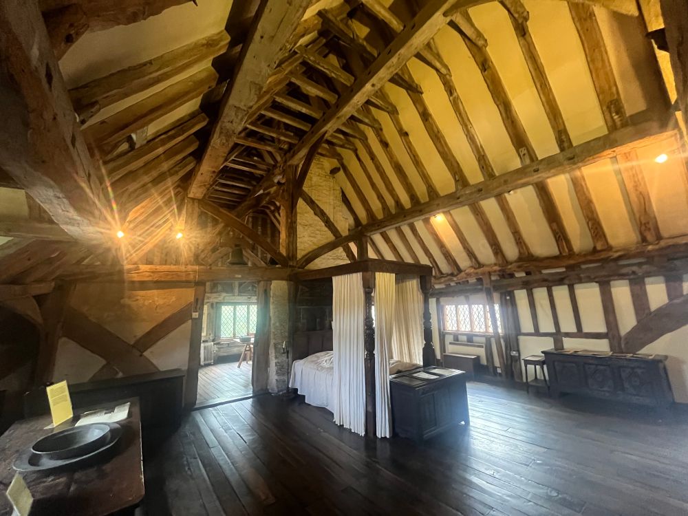 The interior of the bedroom at Anne of Cleves House in Lewes. The building is timber framed with the roof timbers exposed. There is a tester bed in the centre of the space. 