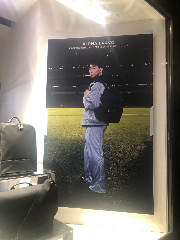 Son wearing a grey tracksuit with a black rucksack, pictured in an advertisement for luggage on a wall (taken in Rockefeller Centre New York in January)