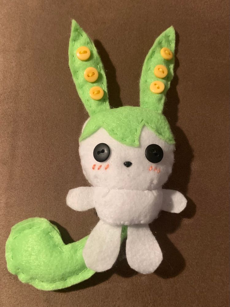 A photo of Zundamon plush front side, made out of felt fabric. Background is in solid brown colour.

It has green rabbit-like ears with 3 yellow buttons sawn vertically making it vaguely resembling green peas.

Top half of head is green while face and rest of it white. It has black buttons as its eyes, a small black triangle nose, and pink cheek blush embroidered as three, short, vertical lines underneath button eyes.

Its torso is short, thick and white. Unlike other parts, arms and legs are made out of singe sheet of felt. Legs are longer than arms and both are white.

Lastly, it has long tail that is green. It is really thick and round towards end of it.