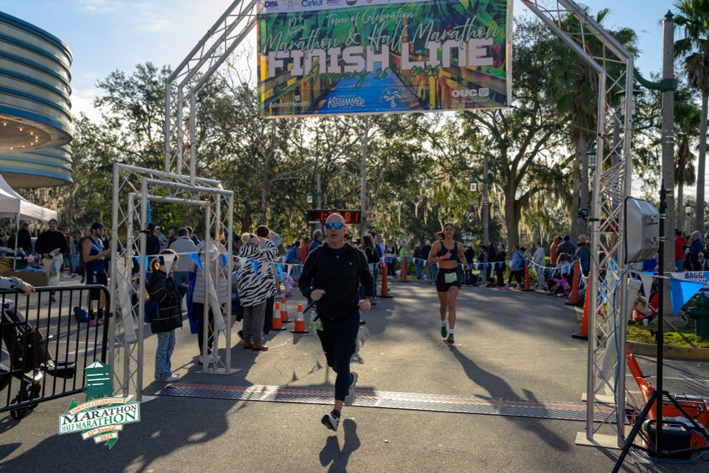 Man running across finish line on a sunny day with people crossing behind. 