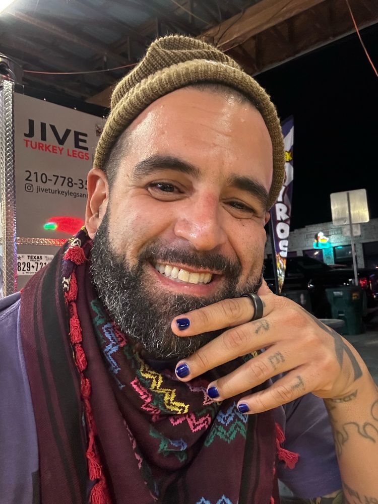 A selfie of Mark. They are smiling and wearing a light brown beanie, a purple shirt, a multi-colored maroon keffiyeh, and their nails are painted sparkly blue. 