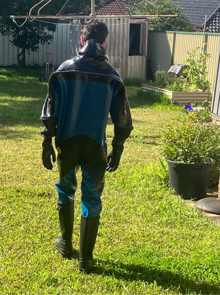 Man walking away from the camera. Wearing a dark blue and black rubber drysuit in the style of Viking brand drysuits. Walking over lawn with plants in the background during the middle of a sunny afternoon.