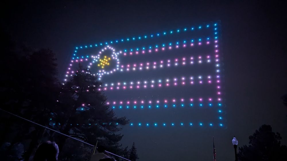 Drones assemble to make a trans Pride flag, with pink, blue, and white stripes, with a sego lily in the upper left corner.