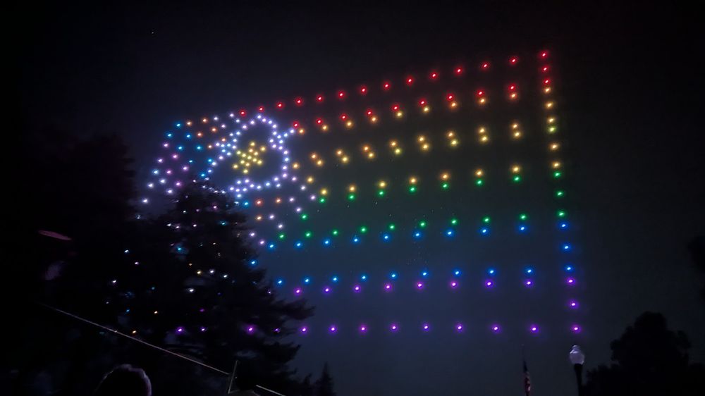Drones in the sky (partly blocked by a tree) assemble Salt Lake City's official Progress Pride flag. It features rainbow-colored stripes, pink and blue triangles on the side, and a sego lily in the upper left corner.