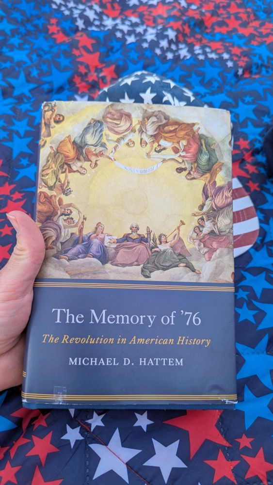 A hand holding the book "The Memory of '76: The Revolution in American History" by Michael D. Hattem. The cover features George Washington surrounded by angels. Behind the book is a baseball cap patterned after the American flag and a blue blanket with red, white, and blue stars.