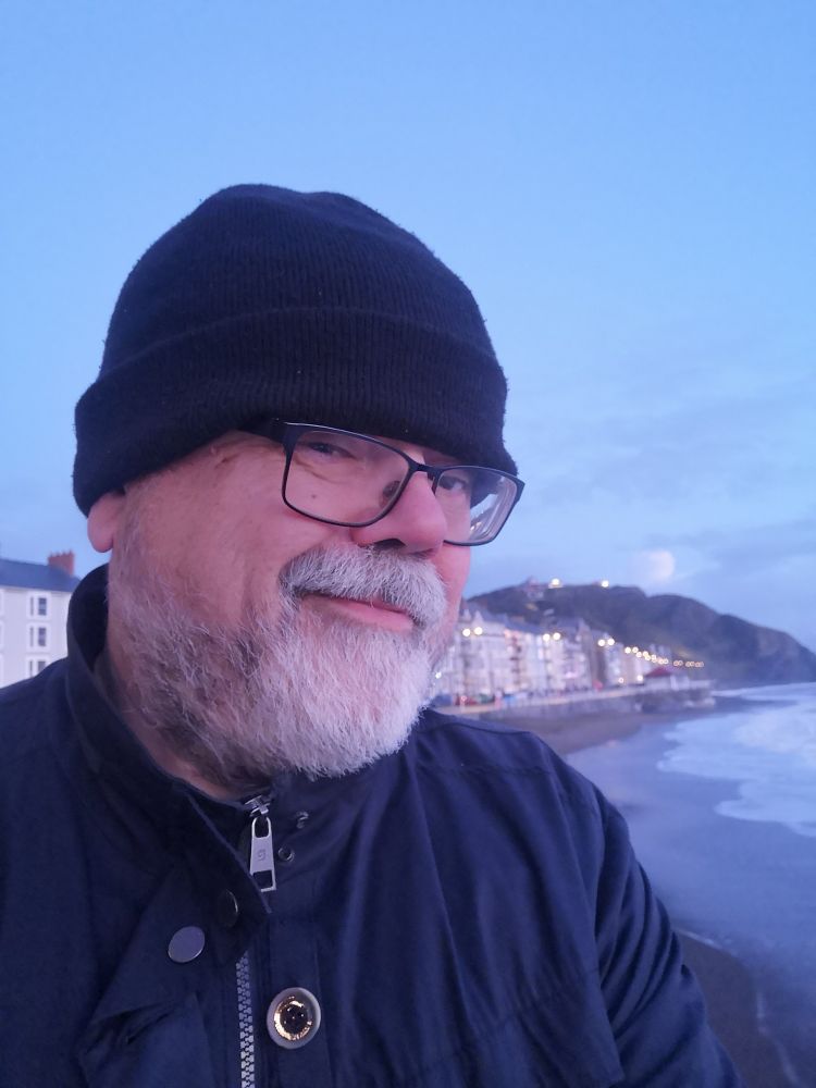 Selfie of well-wrapped-up non-binary person with grey beard on Aberystwyth seafront. Consti (the hill) in background to the north. Waves lapping on the shore.