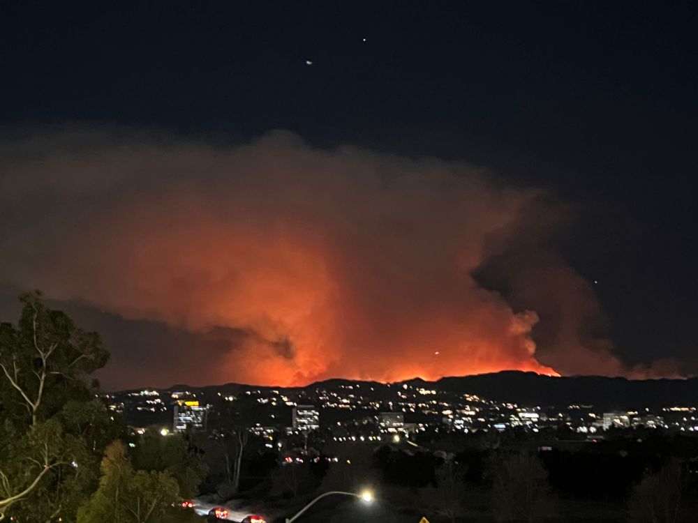 A wide higher-angle shot at night, showing a range of dark hills dotted with lights from houses. At the very bottom of frame the 405 freeway can be seen. Towering over the hills is a dark orange cloud of smoke with fringes of fire at the top of the hills. 