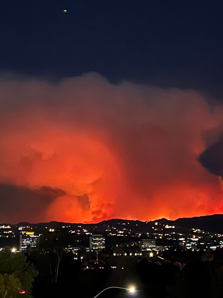 A telephoto higher-angle shot at night, showing a range of dark hills dotted with lights from houses. Towering over the hills is a dark orange cloud of smoke with fringes of fire at the top of the hills. 