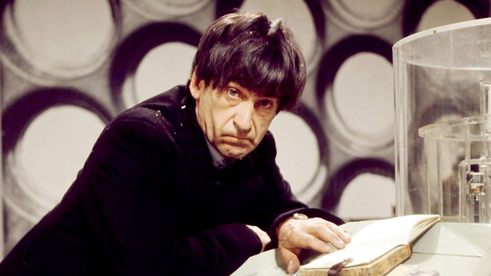 A dark haired man leaning against an electronic control panel. 