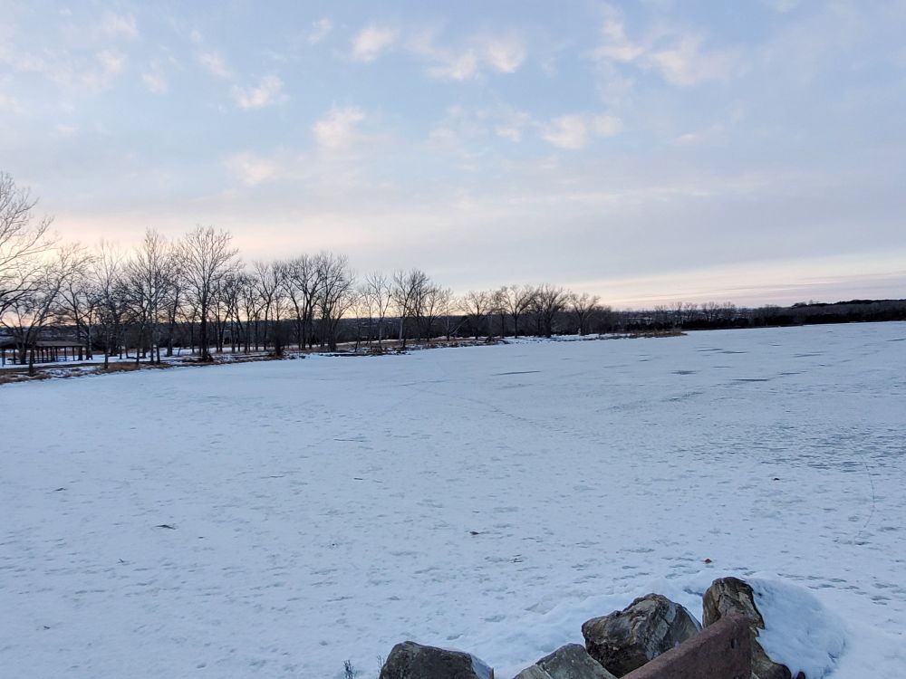 Snow on a frozen overflow pond with trees and a sunset in the background.