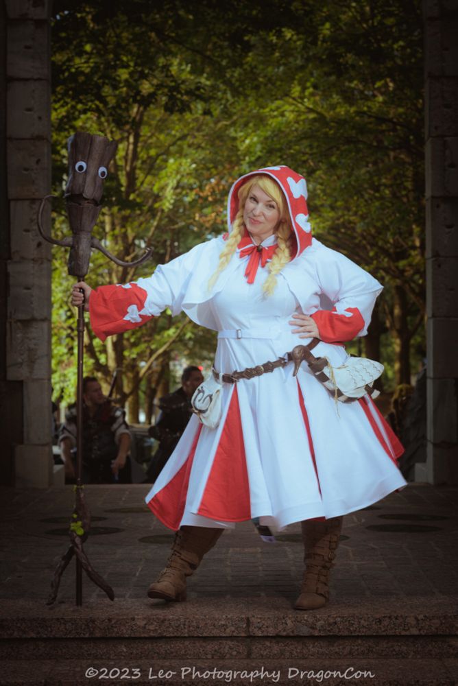 Red’s White Mage cosplay. Cosplay is a white tea length dress with red triangle inserts on a full circle skirt, and a white bolero with wide sleeves abs a hood. The sleeves and hood also have red inserts. Brown belt with a cream pouch on the right hip and a bota bag that looks like a duck on the left hip. Standing leaning on left hip with left hand on hip, right hand holding the Dryad Cane staff. Head tipped to the left, closed mouth smile. Wig is blonde with two braids- one on each side.