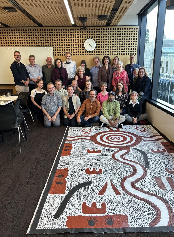 Group of people sitting in front of a large Aboriginal painting. 