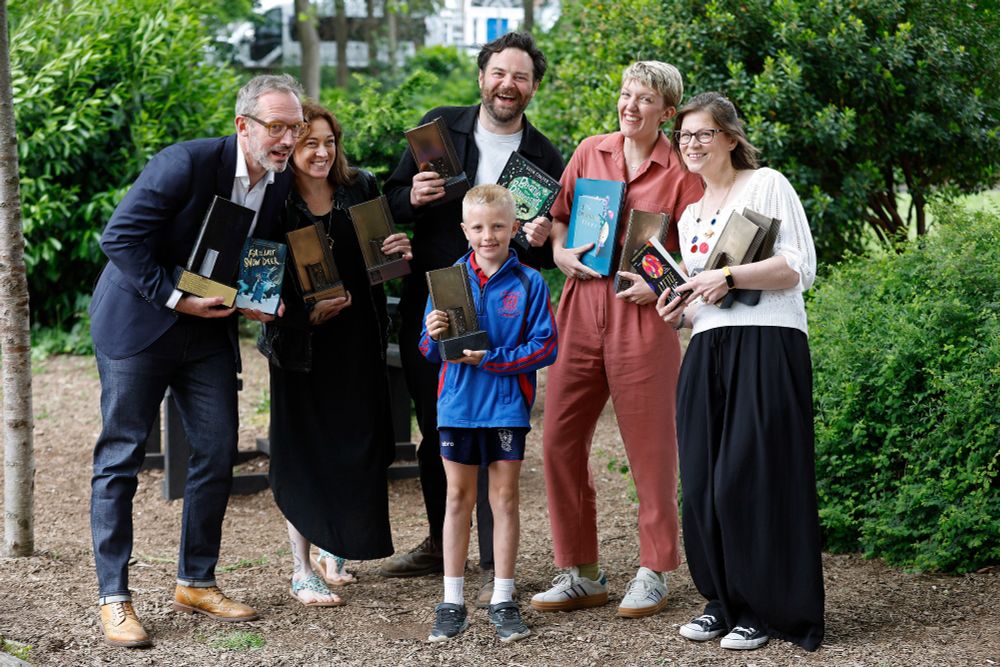 5 adult and one child stand in a clearing with lots of greenery. They are all holding a mixtures of trophys and books and smiling at the camera
